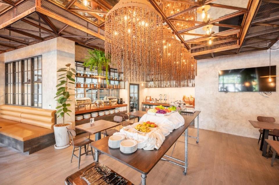 A chic restaurant interior with industrial-style lighting and decor, reminiscent of the stylish dining spaces found in budget hotels near Phuket Airport. A long buffet table, covered in a white tablecloth, holds plates, fruit, and other dishes. Cushioned booth seating lines the left wall beneath exposed beams with hanging lights, while a large TV is mounted on the right wall. Potted plants add a touch of greenery