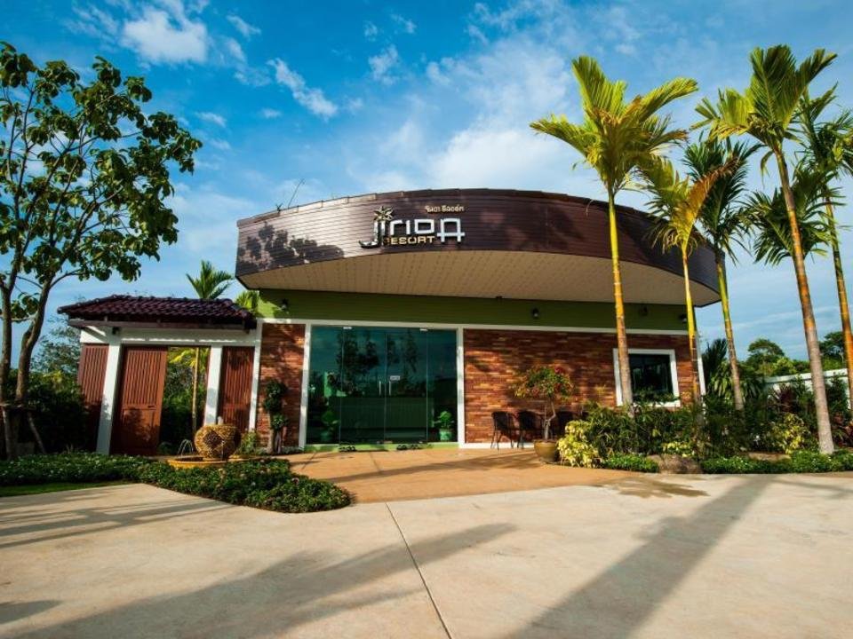 A modern, single-story building with a curved, brown facade labeled "Arima Resort" stands as an elegant option for accommodations near Phuket Airport. It features large glass windows, a covered entrance, and lush greenery including tall palm trees and various plants. The resort is set against a backdrop of blue sky and scattered clouds on a sunny day.