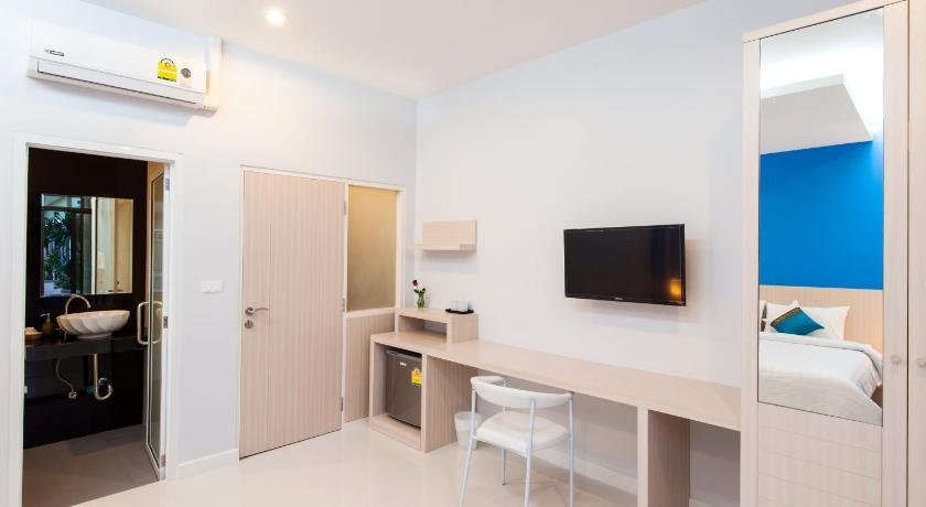 A modern, minimalist hotel room near Phuket Airport. The room features white walls, a mounted flat-screen TV, a long light-colored desk with a white chair, and a small refrigerator underneath the desk. To the left, there is a bathroom with a sink and mirror. A bed with white and blue bedding is visible on the right.