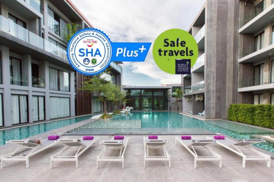 Modern hotel setting with a large swimming pool surrounded by stylish lounge chairs with purple cushions. Two certification logos are displayed: "SHA Plus+" and "Safe travels." The building features balconies overlooking the pool and greenery accents the background, providing a serene ambiance. Perfect for accommodations near Phuket Airport.