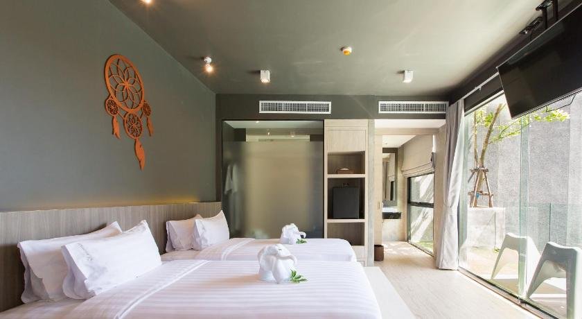 A modern hotel room near Phuket Airport features two neatly made single beds with white linens and towels folded into swan shapes. A dreamcatcher decorates the dark accent wall above the beds. The room includes a flat-screen TV, large windows, and a small bathroom area separated by a frosted glass partition.