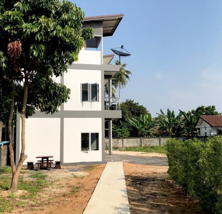 A modern white multi-story house with large windows, a small balcony, and a satellite dish on the roof. A paved walkway leads to the house, with a tree and a wooden picnic table on the left side. The background includes lush greenery, banana trees, and a clear blue sky—ideal accommodations near Phuket Airport.