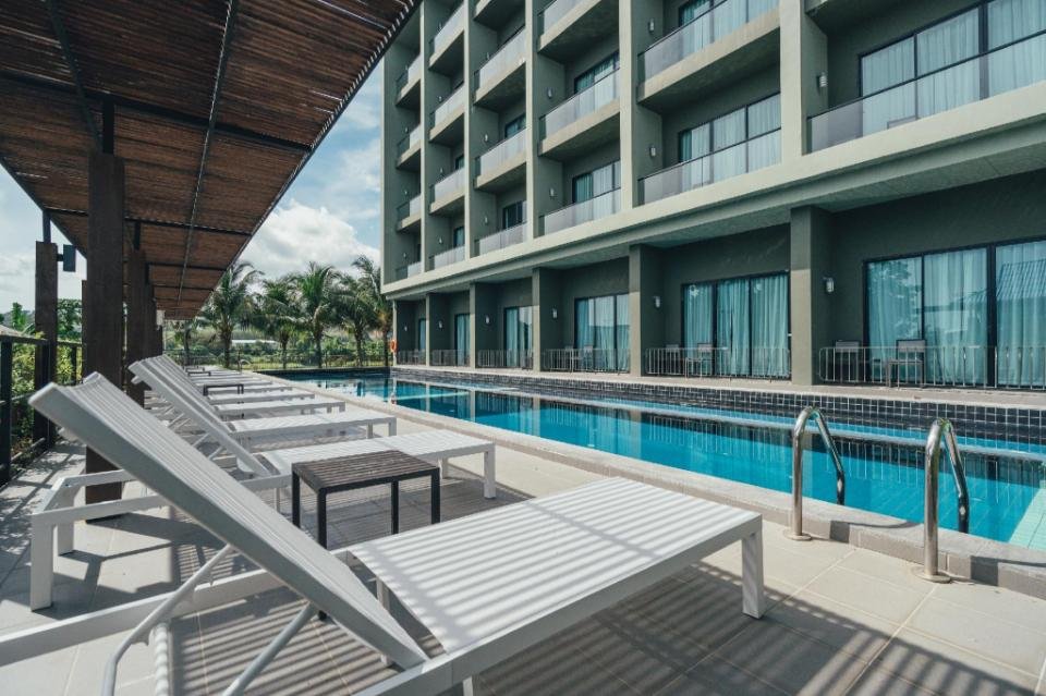 A modern building with multiple balconies overlooks a rectangular outdoor swimming pool lined with white lounge chairs. A shaded area with a wooden canopy runs parallel to the pool. Palm trees can be seen in the background under a partly cloudy sky, typical of budget hotels near Phuket Airport.