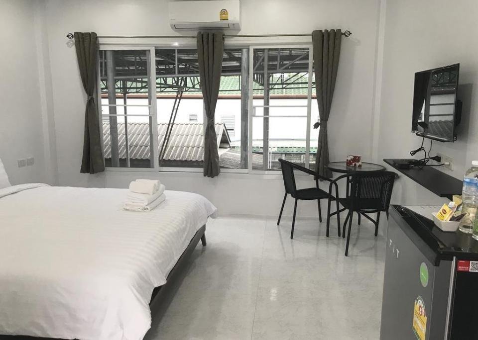 A clean, minimalistic bedroom features a neatly made double bed with white linens and folded towels on top. The room includes a wall-mounted TV, a small round table with two chairs, and a mini-fridge. Three big windows with brown curtains overlook an outdoor structure, ideal for budget hotels near Phuket Airport.