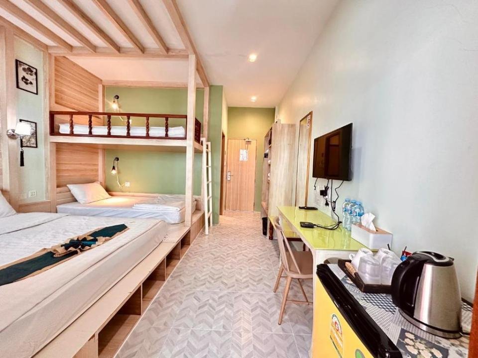 A tidy and bright hotel room featuring a twin bed and a set of bunk beds, located among budget hotels near Phuket Airport. The room includes a wall-mounted flat-screen TV, a yellow mini-fridge, and a desk area with a chair. Adorned with light wood furnishings and green accent walls, essentials like water bottles and a kettle are provided.