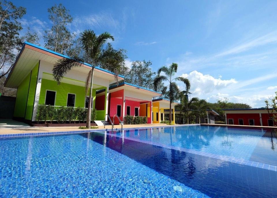 A vibrant resort featuring brightly colored buildings in green, red, yellow, and blue. Each structure has a covered patio with lush greenery and palm trees. In the foreground, a sparkling blue swimming pool reflects the clear sky. Perfect accommodations near Phuket Airport for a serene escape.