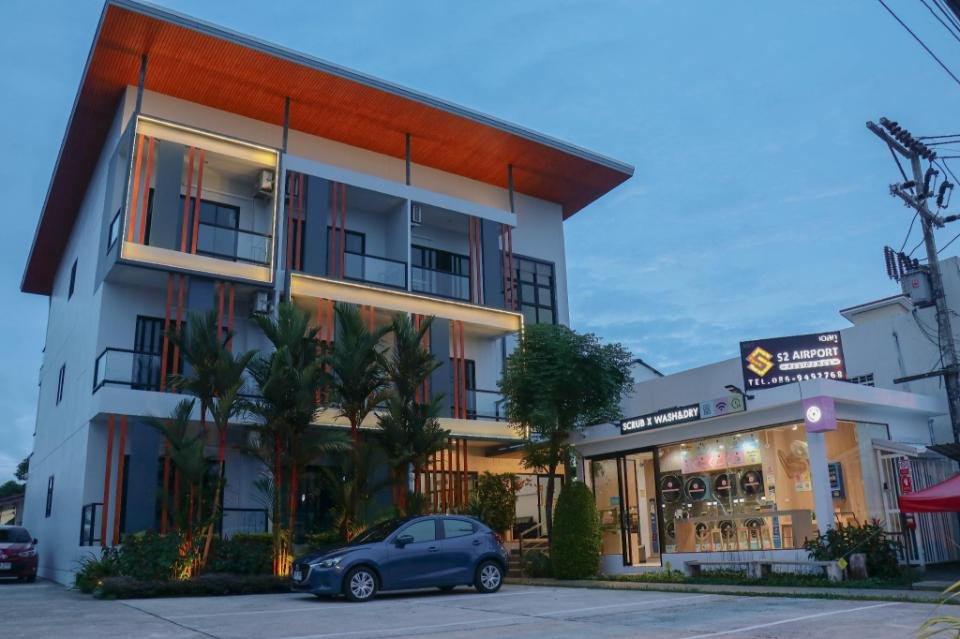 A four-story building with a modern design stands next to a smaller, single-story shop labeled "S2 Airport." The main building, one of the budget hotels near Phuket Airport, has large windows, balconies, and an angled roof. Several palm trees and a parked car are in front, and the surrounding area appears clean and well-maintained.