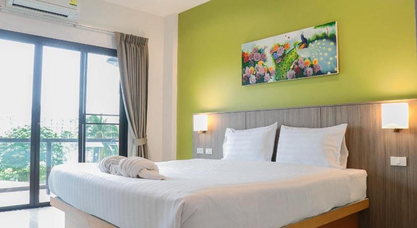 A modern bedroom in a hotel near Phuket International Airport features a large bed with white linens and a folded towel. The green-painted wall behind the bed is adorned with a colorful painting of flowers and a peacock. A sliding glass door with beige curtains leads to a balcony overlooking trees.