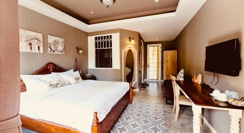 A cozy hotel room with a large wooden bed and white linens. The beige walls complement the window's white trim. A wall-mounted TV, wooden desk with a lamp, tea set, and chair are on the right. The tiled floor patterns enhance the two paintings above the bed. Ideal accommodations near Phuket International Airport.