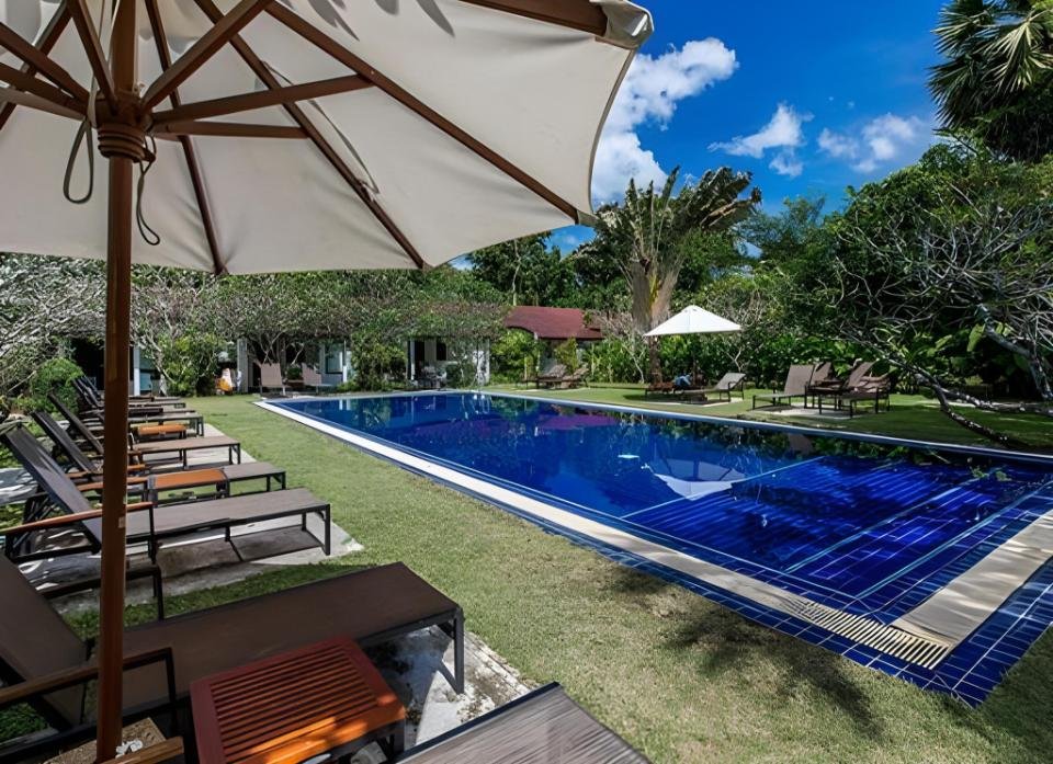 A serene outdoor pool scene with a rectangular blue-tiled swimming pool surrounded by lush greenery at one of the budget hotels near Phuket Airport. Lounge chairs and large umbrellas are arranged neatly by the grassy lawn, while the clear, blue sky provides a bright and sunny atmosphere overall.