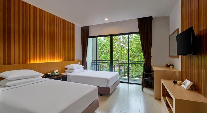 A modern hotel room near Phuket International Airport features two single beds with white bedding, wooden headboards, and matching nightstands. The room has wood-paneled walls, a flat-screen TV, a desk with a mirror, and large windows with curtains, offering a view of lush green trees from a balcony.