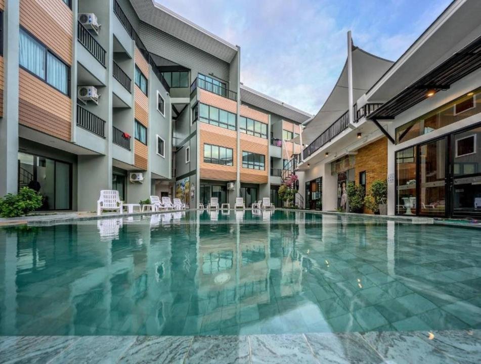 Apartment complex with a large central swimming pool. The modern buildings have three stories and feature balconies and large windows. Poolside chairs are arranged along the edge, and there are arched canopies providing shade. Accommodations near Phuket Airport create a serene atmosphere under a partly cloudy sky.