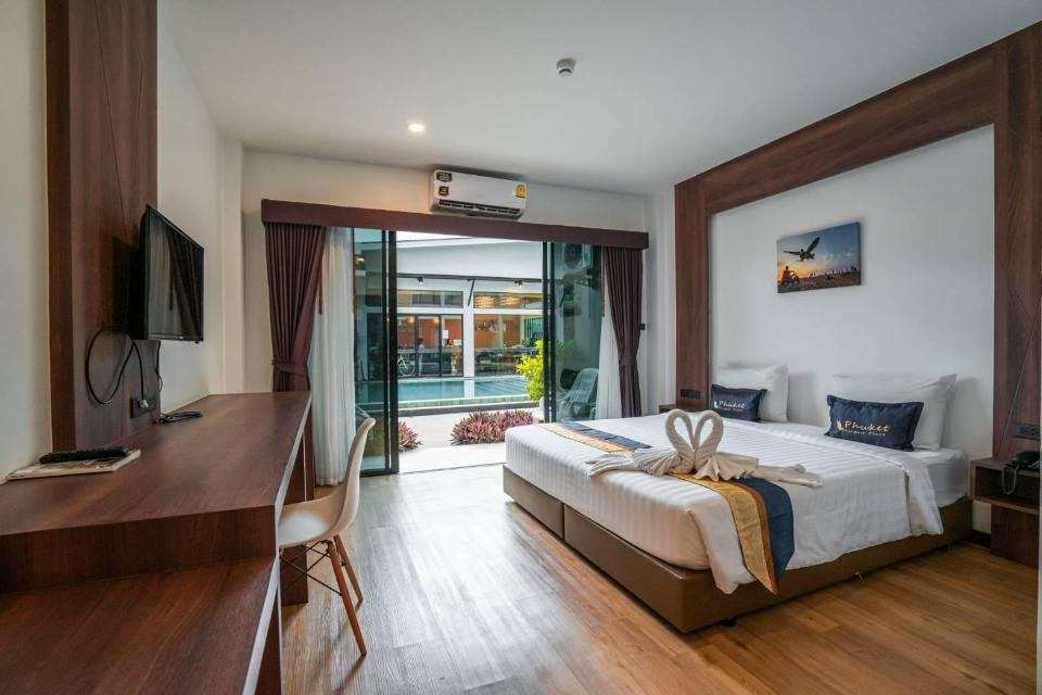 Modern hotel room near Phuket International Airport with hardwood flooring featuring two single beds pushed together. Beds are adorned with swan-shaped towels and blue pillows. A wooden desk with a chair and TV is on the left. Glass doors open to a patio with a garden view, complementing an aviation-themed painting.