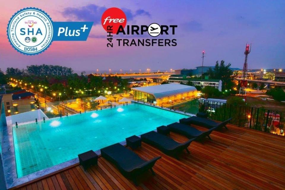 A rooftop pool with an infinity edge overlooks a cityscape at sunset. Four lounge chairs are arranged on the wooden deck beside the pool. The sky is a gradient of purple and orange. Text reads "SHA Plus+ 24HR free AIRPORT TRANSFERS." City lights and buildings are visible in the background, ideal for Accommodations near Phuket Airport.