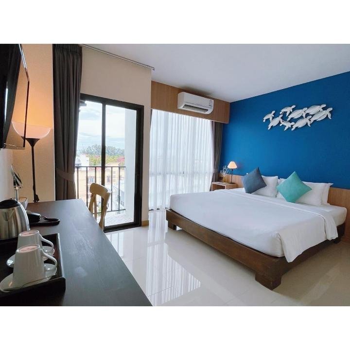 A modern hotel room near Phuket Airport features a large bed with a white duvet and blue and green pillows against a blue accent wall adorned with a fish sculpture. A sliding glass door leads to a balcony with outdoor scenery. The room includes a wooden desk, chair, and coffee station with cups and kettle.