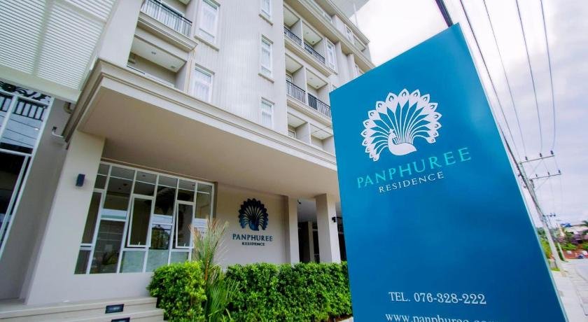 A modern, multi-story building with a simple facade is shown. In the foreground, a large blue sign with a white peacock logo reads "PANPHUREE RESIDENCE," along with a telephone number and a website URL. Ideal for those seeking budget hotels near Phuket Airport, the entrance boasts large windows and a small green garden with various plants.