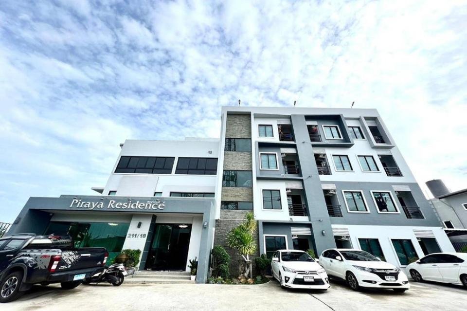 A modern four-story building, "Piraya Residence," features a blend of white, gray, and stone exteriors. Several vehicles including cars and motorcycles are parked in front. The sky above is partly cloudy with blue patches. This property is one of the top accommodations near Phuket Airport.