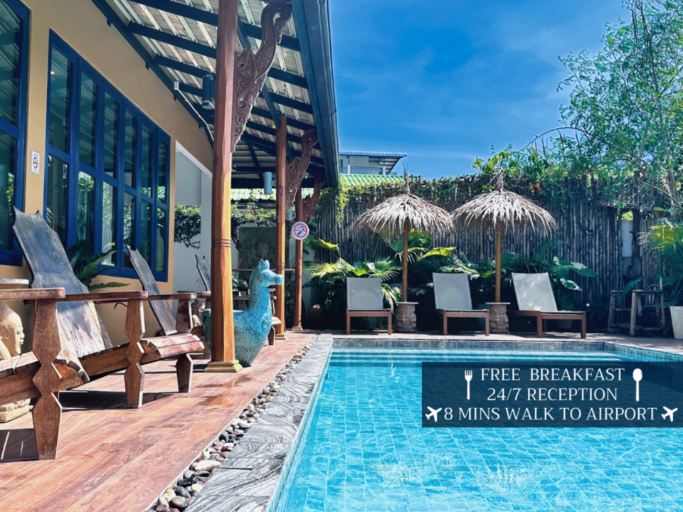 A serene poolside area at this tropical hostel near Phuket Airport features wooden loungers, thatch umbrellas, and lush greenery. Blue-tiled pool waters shimmer under clear skies. A sign reads: "FREE BREAKFAST, 24/7 RECEPTION, 8 MINS WALK TO AIRPORT," highlighting convenient guest services.