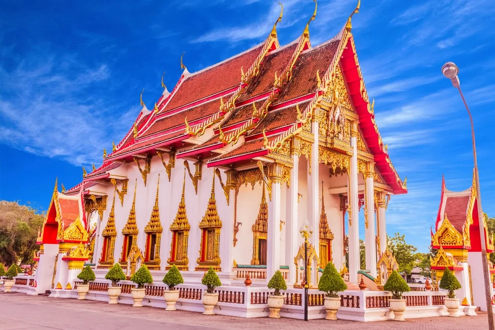 A grand Thai Buddhist Temples in Phuket with ornate gold and red detailing stands under a bright blue sky. The Temples in Phuket features intricate carvings and pointed roofs adorned with elaborate patterns. Lush green shrubs and trees line the pathway leading to the main entrance, enhancing its majestic appearance, much like the luxurious hotels in Phuket nearby.