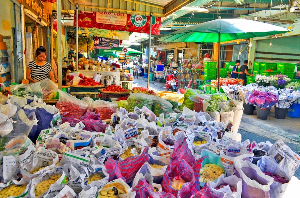 A vibrant outdoor market scene with various colorful goods, including fruits, vegetables, and flowers. Stalls are covered with canopies and parasols, reminiscent of the bustling natural attractions in Bangkok. Shoppers browse through the stalls, where items are packaged in bags and plastic wraps and signs with text are visible overhead.