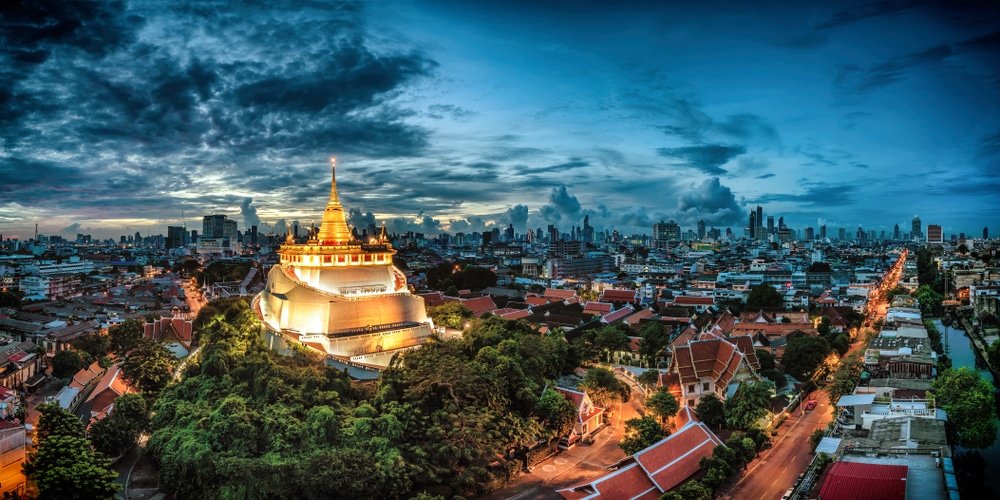 A vibrant panoramic view of Wat Saket, one of the natural attractions in Bangkok, also known as the Golden Mount, at dusk. The illuminated temple stands prominently with its golden spire, surrounded by lush greenery and an urban landscape. The sky is a mix of dark and blue hues with scattered clouds.