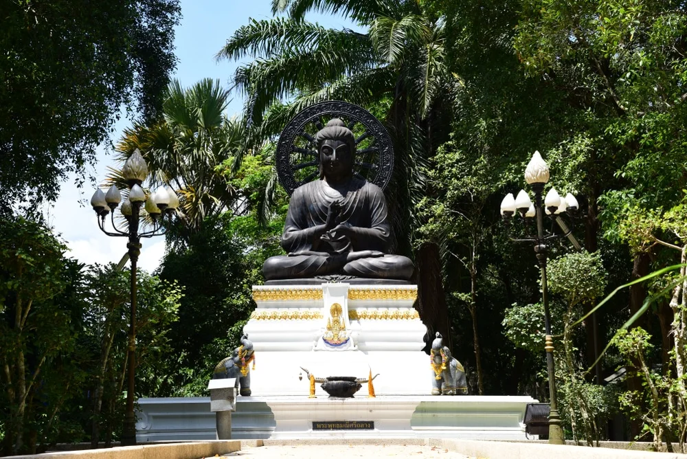 A large statue of a seated Buddha is situated in a lush, green park. Positioned on an elevated white platform adorned with smaller statues and decorations, it creates a serene atmosphere that travelers to the nearby hotels in Phuket often visit. Street lamps and dense trees complete the peaceful setting.