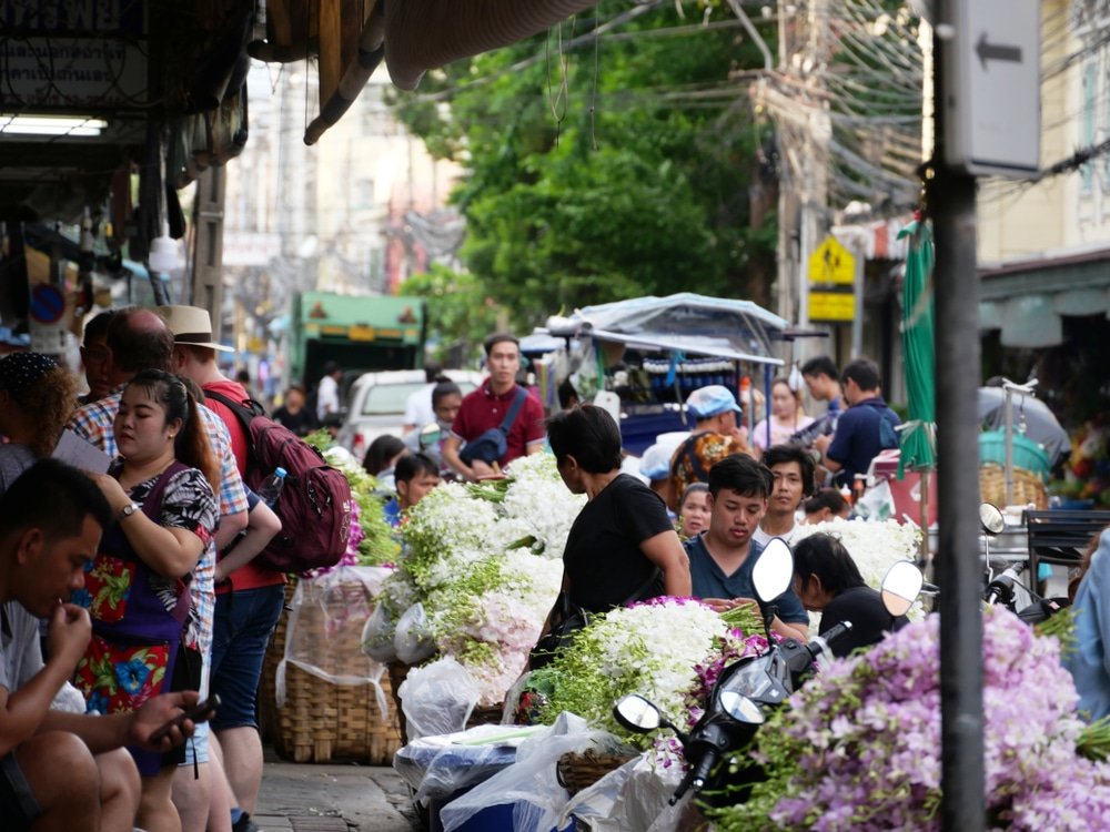 A bustling street market, one of the natural attractions in Bangkok, is filled with vendors and shoppers. Stalls are adorned with various flowers, mainly white and purple. People are engaged in buying and selling. In the background, buildings and hanging wires are visible, adding to the busy atmosphere. Motorcycles are parked nearby.