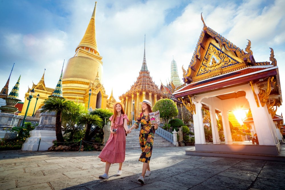 Two women in vibrant dresses and hats walk hand-in-hand, smiling, and holding maps in a grand temple complex. Surrounding them are ornate golden spires and traditional Thai architecture with intricate detailing, emblematic of the natural attractions in Bangkok. The bright sky and sunlight create a warm, welcoming atmosphere.