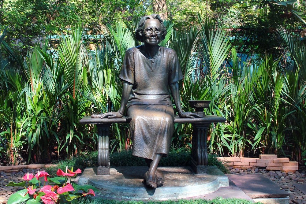 A bronze statue of an elderly woman sitting on a bench is surrounded by lush greenery, reminiscent of the natural attractions in Bangkok. She is wearing glasses, a dress, and has a gentle smile. Her legs are crossed, and her hands rest on the bench. Vibrant red and pink flowers bloom in the foreground.