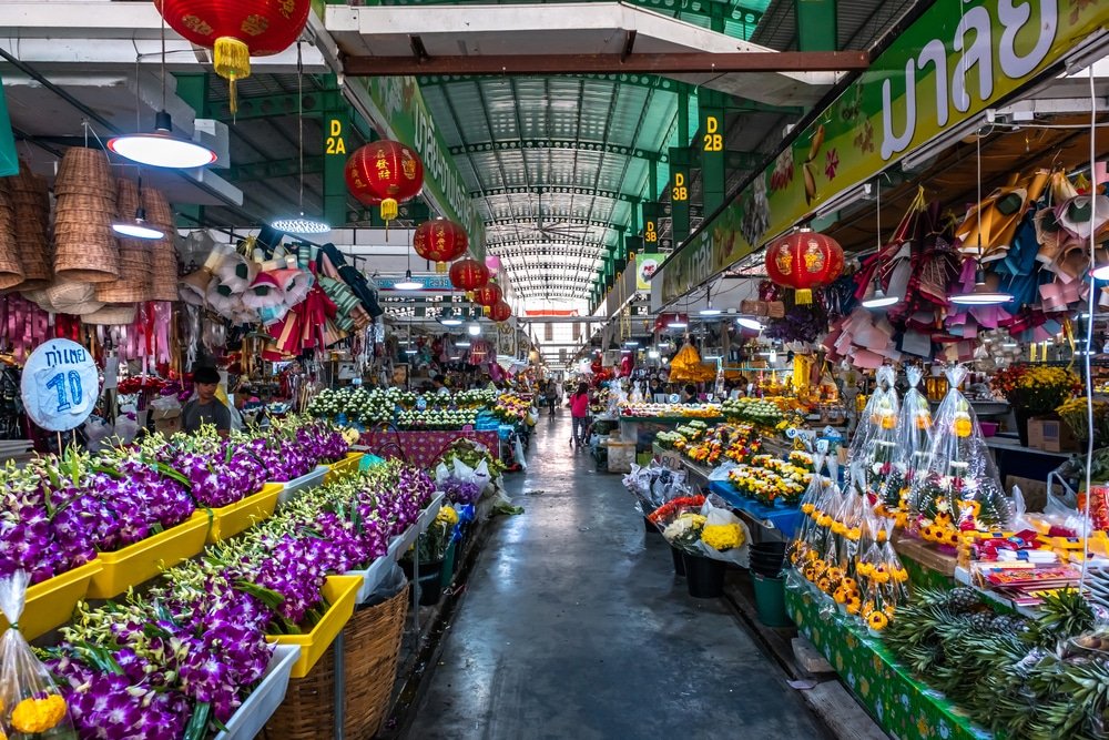 A vibrant indoor market, one of the natural attractions in Bangkok, features stalls with a variety of colorful flowers, plants, and produce. The ceiling is adorned with red lanterns, and signs in a non-Latin script are visible. The bustling market is filled with shoppers, and narrow pathways are lined with bright, fresh blooms and greenery.