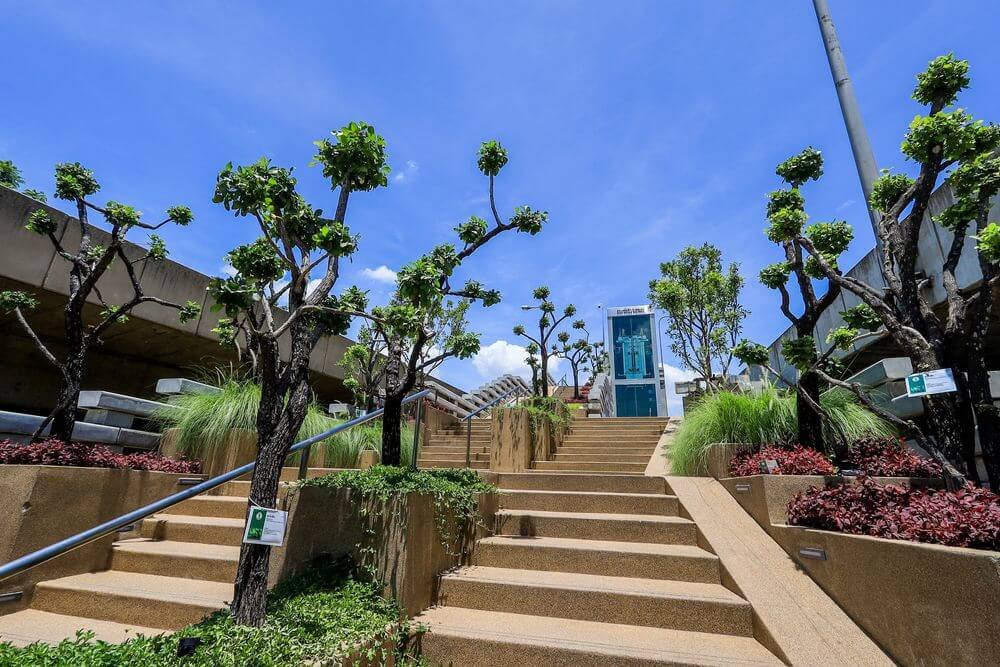 A series of wide, light brown stairs ascend an outdoor area, flanked by neatly pruned trees and lush vegetation, including green grasses and red-leaved plants. A glass elevator shaft stands prominently at the top of the stairs against a backdrop of a blue sky with a few clouds—reminiscent of natural attractions in Bangkok.