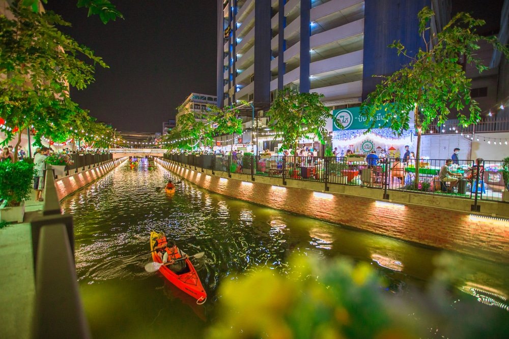 A vibrant night scene features a canal flanked by bright city lights and buildings. People are kayaking in the water, while others dine at outdoor restaurants along the canal's edge. Lush trees and flowers frame the view, adding a natural touch to these Natural Attractions in Bangkok.