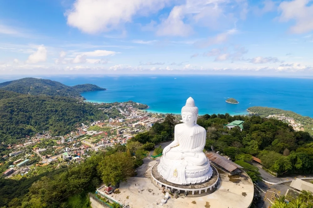 Aerial view of the Big Buddha statue in Phuket, Thailand, with lush green hills in the foreground and turquoise ocean waters in the background. The statue is a large, seated white Buddha overlooking the coastal town's scattered buildings and roads amid the verdant landscape near numerous temples in Phuket.