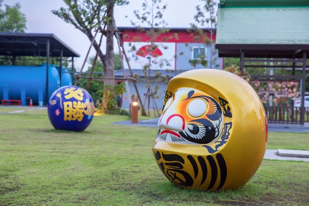 A photo of a grassy outdoor area featuring two large daruma dolls captures the essence of natural attractions in Bangkok. The closer doll is gold with intricate black, red, and white facial designs. In the background is a blue daruma doll adorned with large gold kanji characters, along with trees and buildings creating a park-like setting.