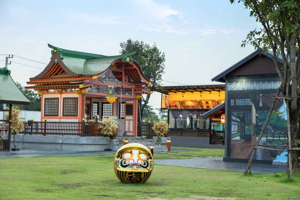 The image features a Japanese-style temple with ornate wooden architecture and green roof tiles. In the foreground, a large golden Daruma doll with intricate designs stands on a grassy area, reminiscent of natural attractions in Bangkok. Trees and additional traditional buildings are visible in the background, with a dusky sky overhead.