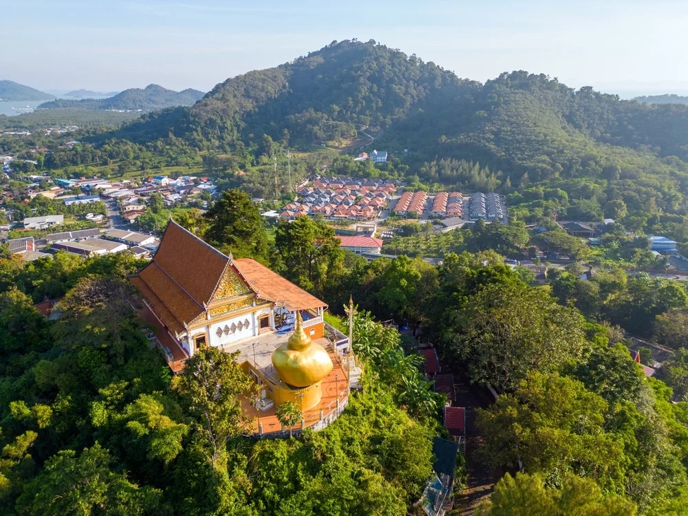 Aerial view of a Buddhist temple with an orange roof, nestled among dense green trees on a hilltop. A large golden Buddha statue is prominent in the foreground. Surrounding the temple, numerous houses and buildings extend towards forested mountains, reminiscent of the tranquil setting found at hotels in Phuket.