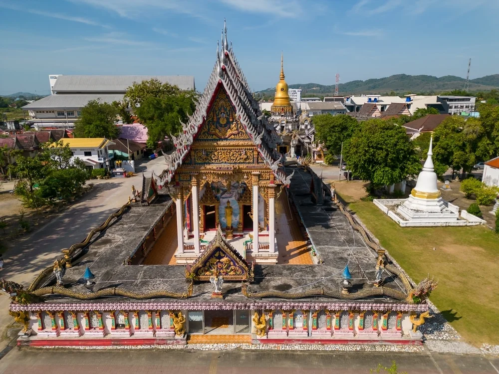 Aerial view of a Thai temple complex featuring intricate, colorful roof architecture and statues across the grounds. The ornate main building has a gabled roof with detailed carvings and figures. Trees, a golden stupa, and other small structures are set against a backdrop of distant hills and hotels in Phuket.