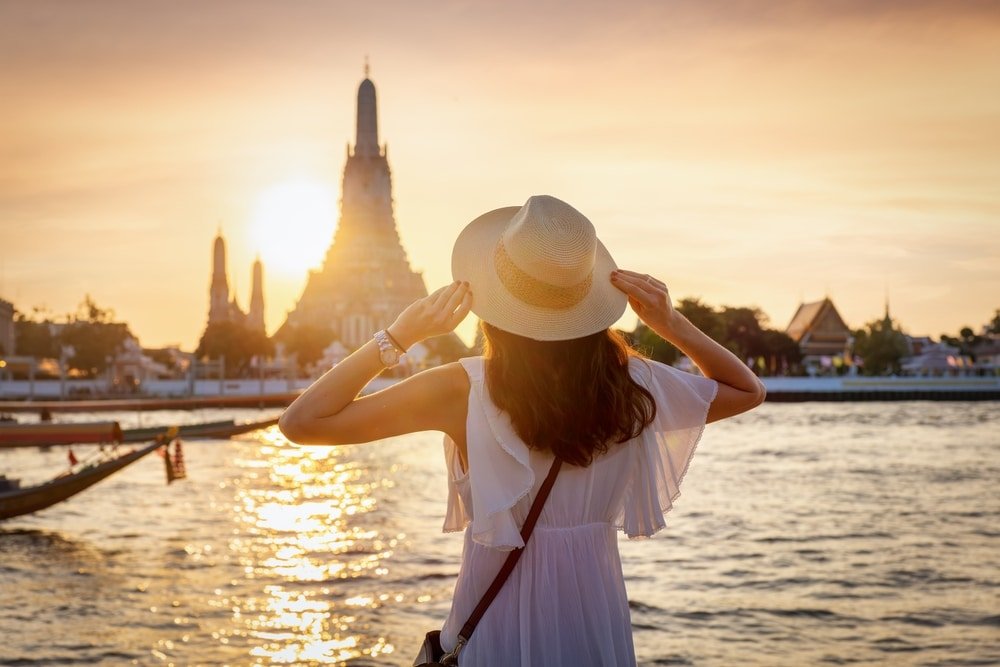 a woman wearing a hat and standing by water with a building in the background