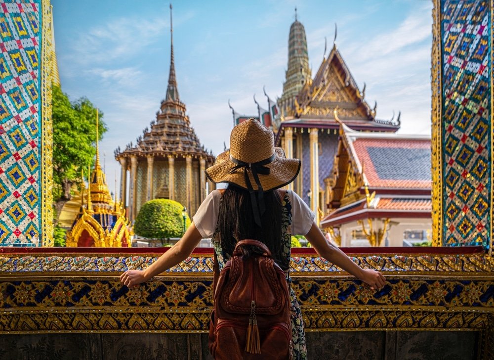 A woman with long dark hair wearing a woven hat and a brown backpack looks out at one of the natural attractions in Bangkok, a colorful, ornate temple complex featuring intricately designed towers and golden structures under a blue sky. She stands framed by a doorway adorned with vibrant mosaic patterns.