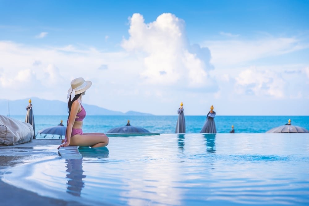 A woman in a pink bikini and wide-brimmed straw hat sits at the edge of an infinity pool, looking out at the ocean. The sky is clear with just a few fluffy clouds. Beach umbrellas are seen in the distance, and hills are visible on the horizon. The scene, typical of hotels in Phuket, is serene and picturesque.