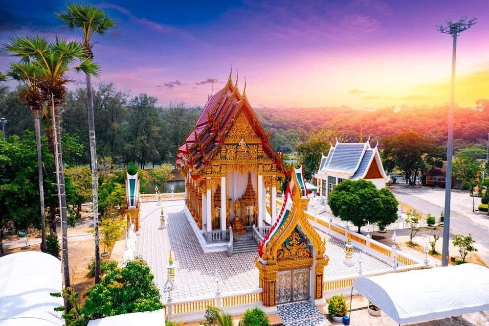 Aerial view of a picturesque Buddhist temple at sunset, featuring ornate golden and red roof structures and intricate architectural details. Nestled among lush greenery and tall palm trees, this gem, one of the renowned temples in Phuket, basks under a vibrant sky showcasing hues of purple, orange, and pink.