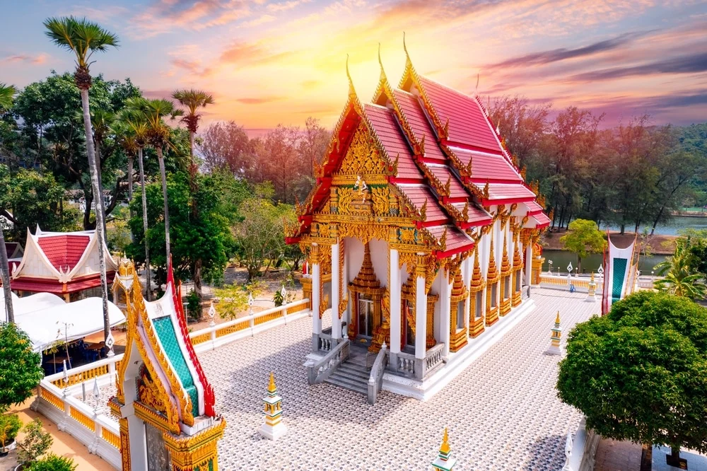 Aerial view of a beautifully ornate Buddhist temple with a red and gold roof, set against a backdrop of a vibrant sunset. The temple is surrounded by lush greenery and other smaller structures. Palm trees and a forested area are visible in the background. The temple grounds, much like the exquisite temples in Phuket, are paved with patterned tiles.