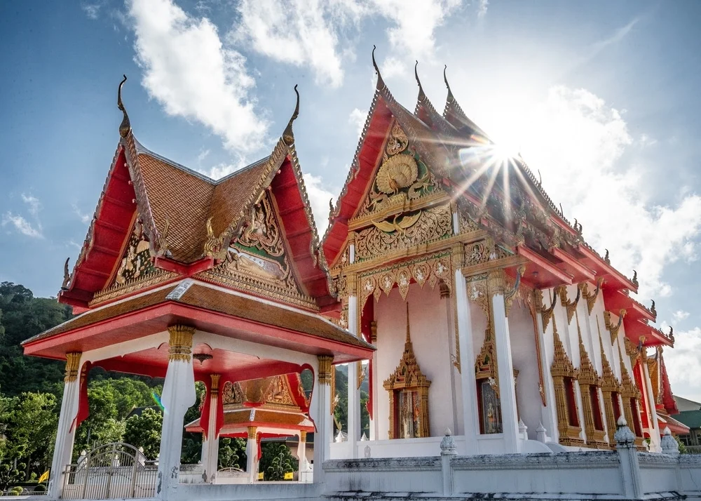 Traditional Thai temple with ornate gold carvings and red roofs gleams under the sunlight. The main worship hall, adorned with intricate artwork, stands beside a smaller pavilion. Bright blue sky with fluffy clouds adds a serene backdrop, while greenery surrounds the structure, akin to hotels in Phuket.