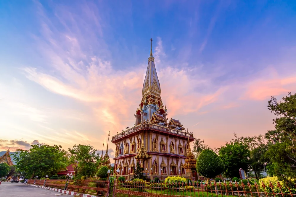 The image shows a stunning temple with intricate architecture at sunset. The sky behind the temple is filled with vibrant colors, ranging from soft pinks to rich purples. Surrounding the temple are lush green trees and neatly maintained gardens, adding to the serene atmosphere similar to Temples in Phuket.