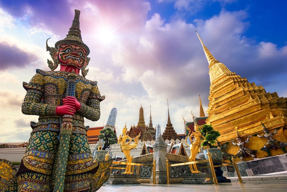 A vibrant scene of Wat Phra Kaew, one of the premier natural attractions in Bangkok, featuring a large, colorful guardian statue with intricate details. The golden pagoda glistens under a partly cloudy sky. Ornate temple structures and mythical statues enhance the grandeur, with sunlight illuminating the complex.