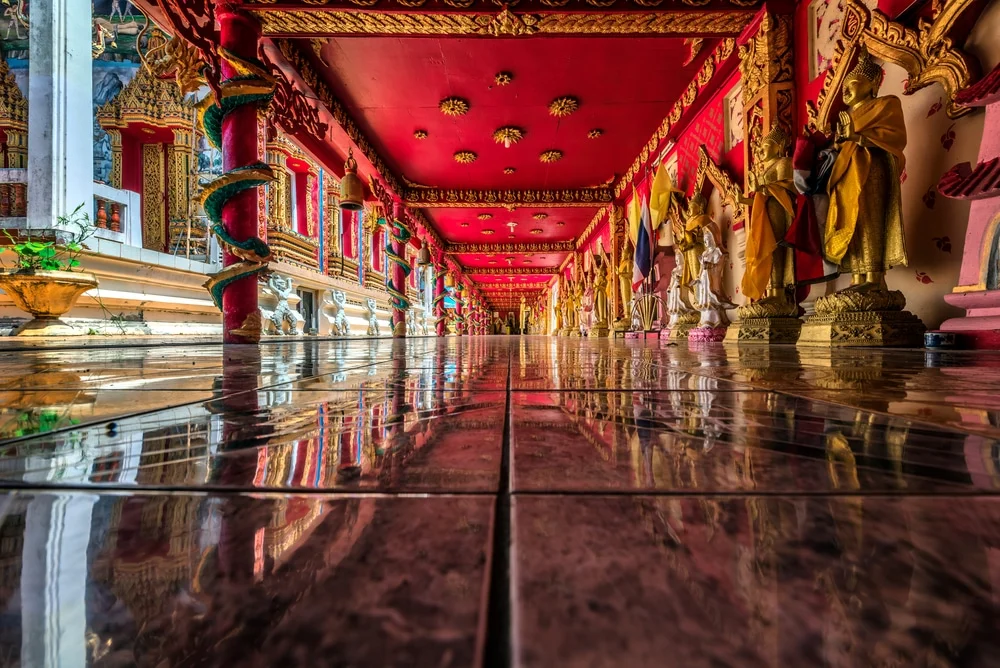 A vibrant temple corridor with a reflective tiled floor leads to a distant exit. The ceiling and columns are adorned with intricate red and gold decorations. Statues and ornate designs line both sides of the corridor, creating a symmetrical and colorful pathway reminiscent of the luxurious hotels in Phuket. Sunlight filters into the space.