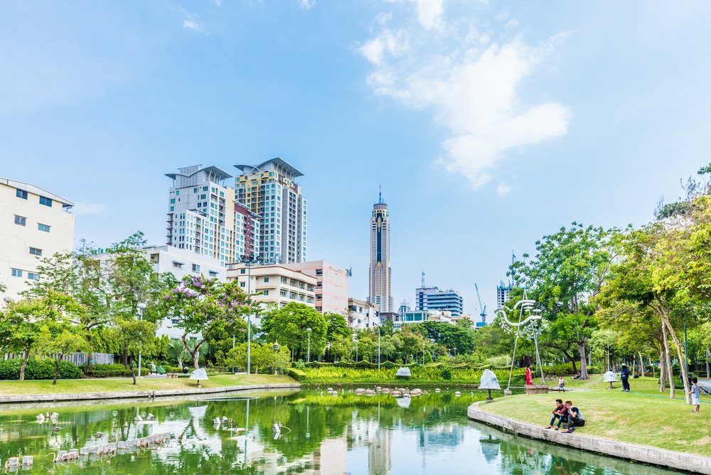 A vibrant city park with colorful flowers, lush greenery, and a tranquil pond takes up the foreground. Modern high-rise buildings tower in the background, under a clear blue sky. Trees and well-maintained gardens surround the area, providing a peaceful contrast to the urban skyline—a perfect example of natural attractions in Bangkok.