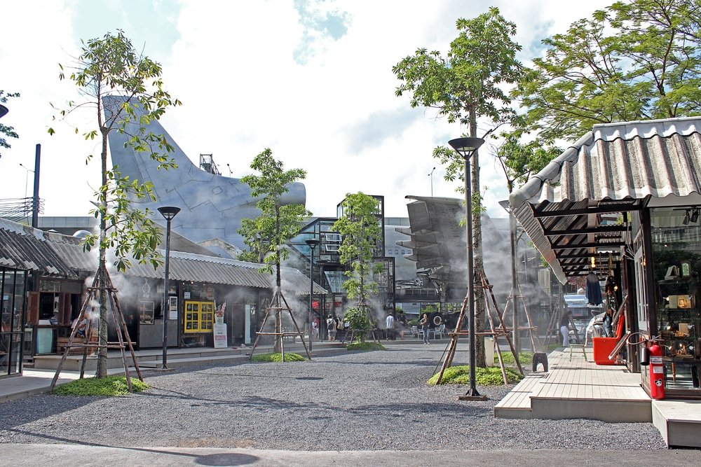 Outdoor shopping area with modern industrial design. It features small shops, greenery, gravel paths, and misting systems to cool the air. There's a large aircraft on display in the background, adding a unique touch. Trees and lamp posts line the pedestrian-friendly walkway—one of the natural attractions in Bangkok.