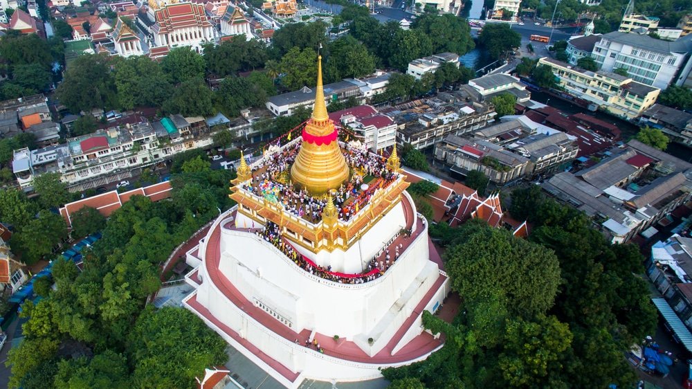 Aerial view of the Golden Mount Temple in Bangkok, Thailand. A large crowd of people gathers on the temple's golden stupa adorned with red, gold, white, and dark accents. Surrounding this natural attraction are lush trees, traditional buildings, and a blend of historic structures with modern architecture.