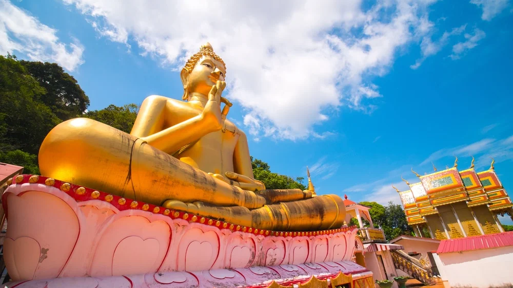 A large golden Buddha statue sits serenely on a lotus pedestal under a blue sky with scattered clouds, resembling the tranquil scene depicted in shutterstock_751429492. The statue's right hand is raised in a meditative gesture. Surrounding the statue are temple structures, including a golden pagoda. Trees can be seen in the background.